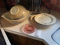 Overview of the set showing nested Pyrex mixing bowls, two dinner plates, and pink glass saucers on a countertop.