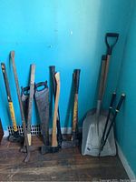 Full view of shovels, saws, axes, pry bar and sledgehammer leaning against a blue wall on wood floor.