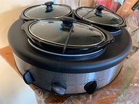 Top view showing three black ceramic pots with glass lids on a round black stainless steel slow cooker base with three control knobs on front.
