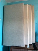 Four stacked rectangular fabric-covered sound panels leaning against a wall, color light beige/gray.