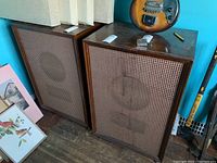 Pair of vintage Lafayette speakers with wooden cabinets and brown grille cloth, placed side by side indoors with a guitar hanging behind one.