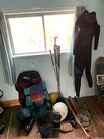 Full view of mixed sports equipment by wall, including multiple bats, helmets, rackets, backpacks, gloves, ball, and wetsuit.