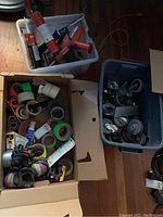 Large cardboard boxes with numerous rolls of different tapes and caulking guns in a plastic bin alongside caster wheels in another plastic bin on a wooden floor.