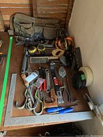 Photo of canvas tool belt and assorted hand tools including wrenches, saw, tape measures, brush, chalk line reels, and more laid out on wooden surface.