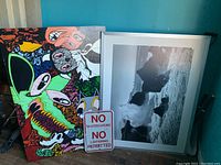 Photo shows colorful abstract face painting, black-and-white ocean wave photograph in white frame, and metal No Skateboarding sign leaning against wall.