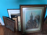 Multiple framed wall art pieces leaning against a turquoise wall, one prominently shows a church artwork with heavy discoloration.