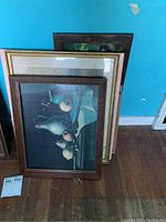 Photo showing four stacked framed art pieces with the top one being a dark still life painting featuring vases and fruit in a brown wooden frame.