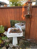 Weathered white pedestal sink used as a planter with dried plants inside, placed near a wooden fence with a large mirror behind it, reflecting the sink.