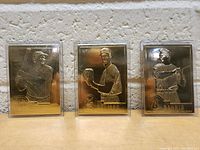 Three gold-colored metal Danbury Mint baseball cards featuring Babe Ruth, Lou Gehrig, and Jim Palmer standing upright against a brick wall.