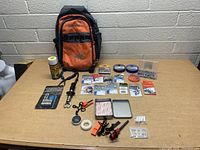 Photo of orange Kilimanjaro backpack with assorted fishing and survival gear spread on table, including fishing line spools, packaged hooks and lures, compass, whistle, and fire starters.
