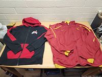 Both jackets side by side on a wooden table; left is black and red Boys Jordan Nike hoodie, right is red and yellow Mens USC Nike jacket.