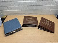 Three stamp albums on a wooden table: two brown Golden Pelican United States Stamps albums and one blue binder.