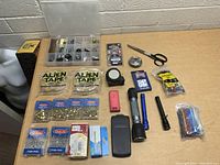 Lot of assorted tools and home items on a wooden surface with Alien Tape, measuring tapes, a compass, scissors, flashlights, USB lighter, packs of push pins, and electricity wire connectors.