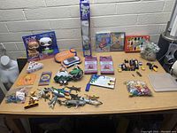 Wide shot of assorted toys on table, showing various toy figures, plush, games, vehicles, and craft items.