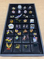 Tray with 34 assorted lapel pins, showing various symbols and designs, organized in a black plastic tray with compartments.