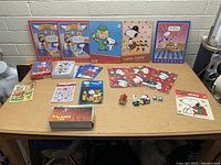 Wide shot showing entire lot of Peanuts Snoopy items including posters, cards, books, figurines, and a balance toy box arranged on a table.