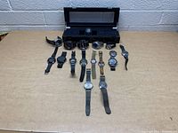 Full collection of 14 watches arranged on a table with a black watch storage case behind them. Shows different styles and bands.