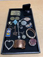 Top-down view of various small items arranged on a black tray including pins, rings, pendants, brooches, bracelets, and a display box with a teardrop amethyst pendant.