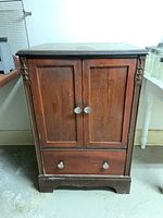 Front view of the vintage wooden cabinet showing two doors and single drawer with metal knobs and handles, placed in a basement area.