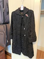 Full view of the black vintage Persian lamb coat hanging on a hook, featuring the small brooch pinned on the chest area.