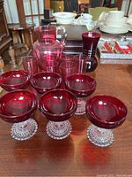 Photo of cranberry glass jug, 6 cranberry glasses, ruby vase and 6 ruby sherbet glasses arranged on table.