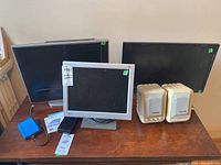 View of entire lot on wooden table showing three monitors of varying sizes, two external hard drives, and a pair of beige speakers.