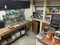 Wide view of workshop corner showing workbenches with numerous plastic containers of assorted hardware items, tools, and plumbing supplies, plus cans on shelves.
