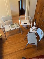 Photo of three shower chairs/supports arranged on a wooden floor in a hallway: one bench-style chair with padded seats and backrest, a composter-style chair with armrests and white toilet seat insert, and a blue plastic shower stool. Bathroom scale and white stool partially visible.