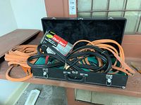 Photo of three jumper cables coiled inside a black carrying case with two silver latches and handle. Cables are thick heavy duty types, orange and black.