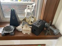 View of entire lot on windowsill showing metronome, vintage camera, brass French horn, miniature piano, Eiffel Tower, two rocks, ashtray, and dumbbell-shaped object.
