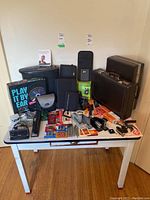 Assorted office supplies and tools arranged on a white table against a cream wall. Items include two briefcases, organizers, staplers, batteries, hole punch, paper shredder, alarm clock, pliers, small tools, and other accessories.