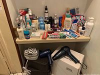 Wide view of all grooming and skincare products on a white shelf above a brown wire basket containing a blue hair dryer, a black Vidal Sassoon hair dryer, and a white box with a Conair set next to it.
