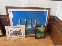 Three framed art prints: large sailing ship print in medium brown frame, medium town scene print in light frame, small rustic cabin painting in dark frame