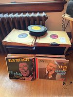 Photo showing stacks of vintage 45s, 78s, and LP albums near a window and radiator, with record sleeves and covers visible.