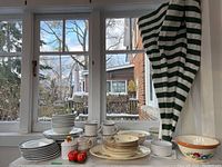 Wide photo showing the full lot on a table by a window, includes plates, bowls, mugs, serving dishes, salt and pepper shakers, and patio umbrella.
