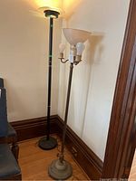 Photo showing two standing lamps side by side against a wall on hardwood floor, one vintage with multiple bulbs and glass shade, one torchiere style with black pole.
