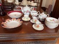 Full view of tea set with teapot, sugar bowl, demitasse cups and saucers, soup bowls, and various plates on carved wooden table.