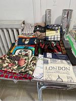 Photo showing multiple folded tea towels and dish cloths with various patterns and colors stacked on a table along with red handled sheathed cutting utensils and kitchen tools in the background. Rolls of aluminum foil and waxed paper are visible standing upright behind the towels.