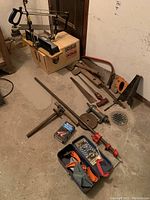 Various clamps, handsaws, an axe head, metal rods, and a boxed mitre saw case on floor against wall