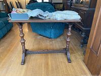 Front view of the wood trestle table with linens and fabric pieces on top showing the legs and carved stretcher.