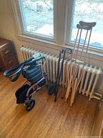 Photo showing the rolling walker, wooden crutches, and wooden canes lined up upright by a window
