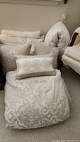 Photo showing comforter, multiple decorative pillows, and body pillow arranged on floor and chair.