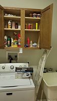 Oak cabinet above washer with various cleaning supplies, Black & Decker iron box on top of washer, metal ironing board leaning against washer.