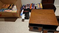 Wide view showing wooden VHS storage case, DVD cases, and multiple boxes of VHS tapes on carpeted floor