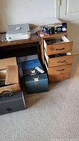 View of wooden drawers with two open drawers revealing miscellaneous office supplies; plastic binders boxed on desk; printer on desk; several file boxes on floor.