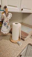 Paper towel holder with wooden base holding new roll of paper towels, next to two beige dish drying mats and glass cutting board on kitchen counter