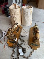 Pair of electric wood wall sconces with metal decorative cutouts and chains, with plaster mounts showing wear. Two cylindrical fabric lamp shades wrapped in paper behind the sconces.