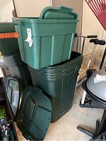 Green plastic trash can with lid and smaller storage tub stacked on top, with some underlying lids visible, next to various garage items.