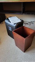 Photo of the black Staples GIBC Shredmaster 50S shredder with the user manual on top and the square wood trash can next to it.