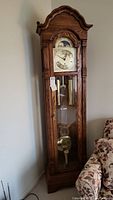 Full view of tall oak grandfather clock showing carved top and glass front door with pendulum and weights inside.